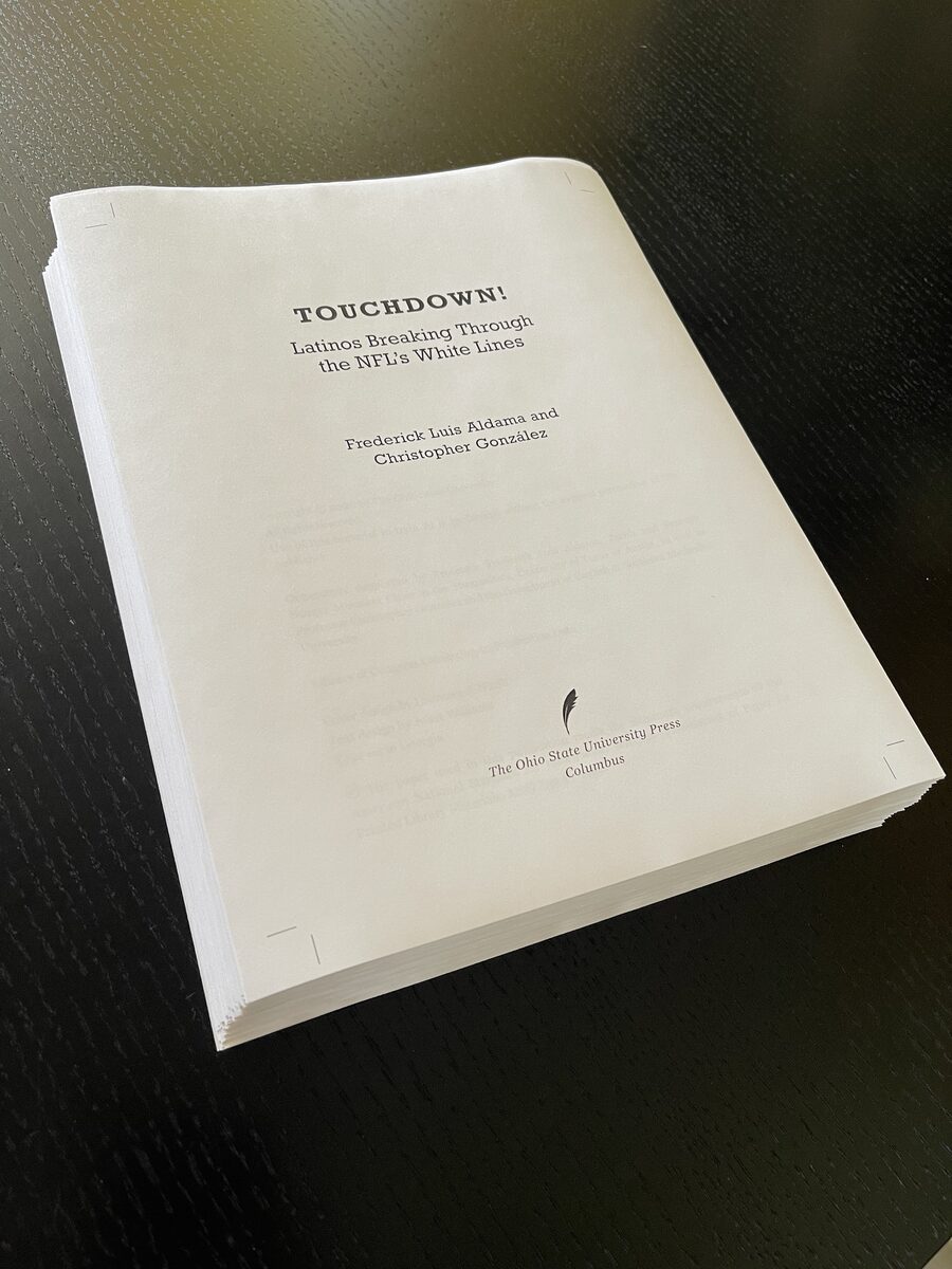 A thick stack of printed page proofs on a dark table; the title page reads Touchdown! Latinos Breaking Through the NFL's White Lines, by Frederick Luis Aldama and Christopher González, The Ohio State University Press